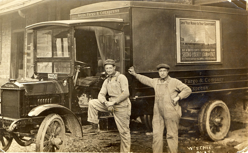 Two men pose in front of an early model Federal motor truck. One man stands with one foot on running board, drivers side. Second man leans with right hand against canvas topped bed of truck. On canvas top side is a square poster showing the U.S. Treasury building in Washington and words Lend money to your government buy a United States government bond Second Liberty Loan of 1917.