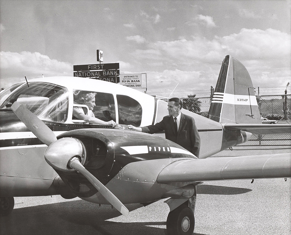 A man in suit and tie stands behind the left wing of a small twin engine plane. He is reaching up to receive a bank passbook being handed out a small window in the plane cockpit by a woman. She sits in the plane’s pilot seat. Signs on chain-link fence and tarmac in background read First National Bank of Arizona and Entrance to nation’s first fly in bank.