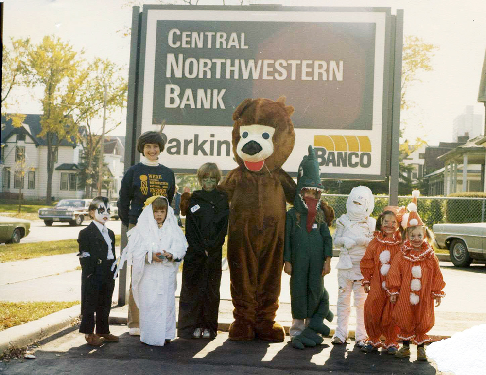 Person in bear costume poses with seven costumed children and one other non-costumed adult in front of sign reading Central Northwestern Bank.