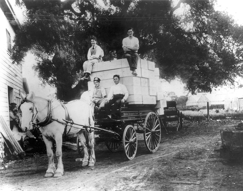 Two men seated on the driver's seat of a wagon with the words Wells Fargo painted on the side. In the bed of the wagon, two other men sit on stacks of white boxes. A white horse stands ready to pull the wagon. Black and white image.