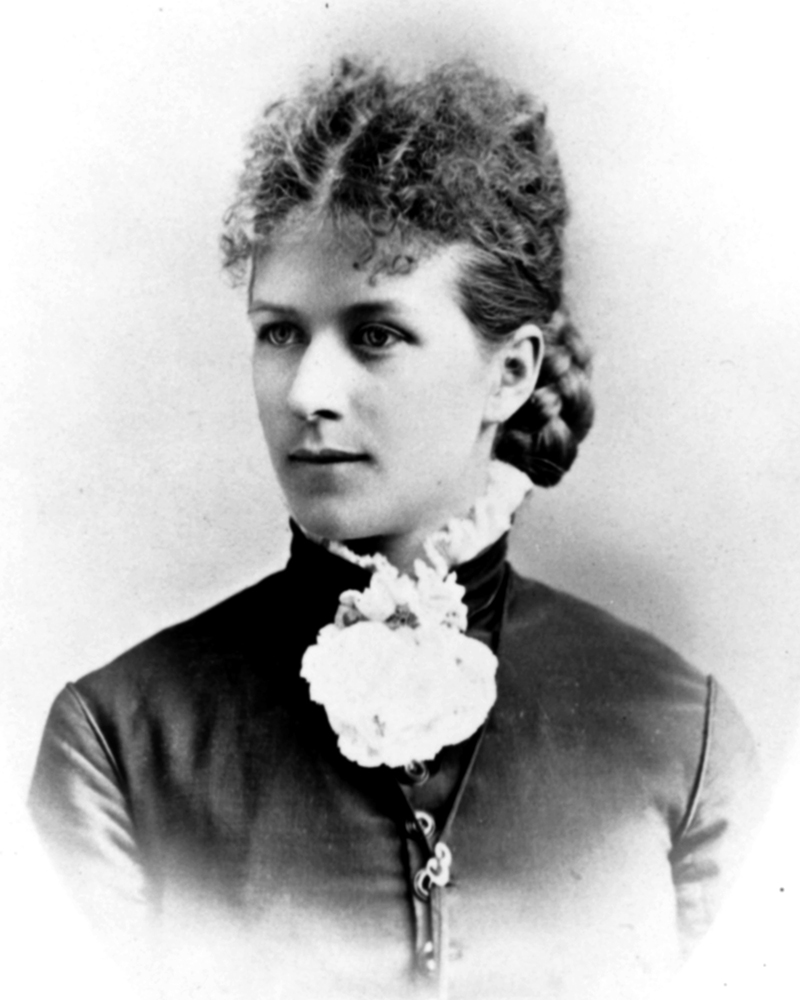 A woman wearing black coat and high ruffle collared blouse with hair pulled back. Black and white portrait.