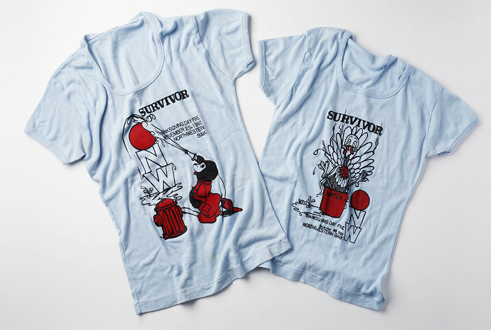 Two blue t-shirts. One features a teddy bear in a red fire fighter suit using a fire hose to put out a fire. One features a turkey in a red water bucket. Both contain words: Survivor, Thanksgiving Day Fire November 25, 1982, Northwestern National Bank.