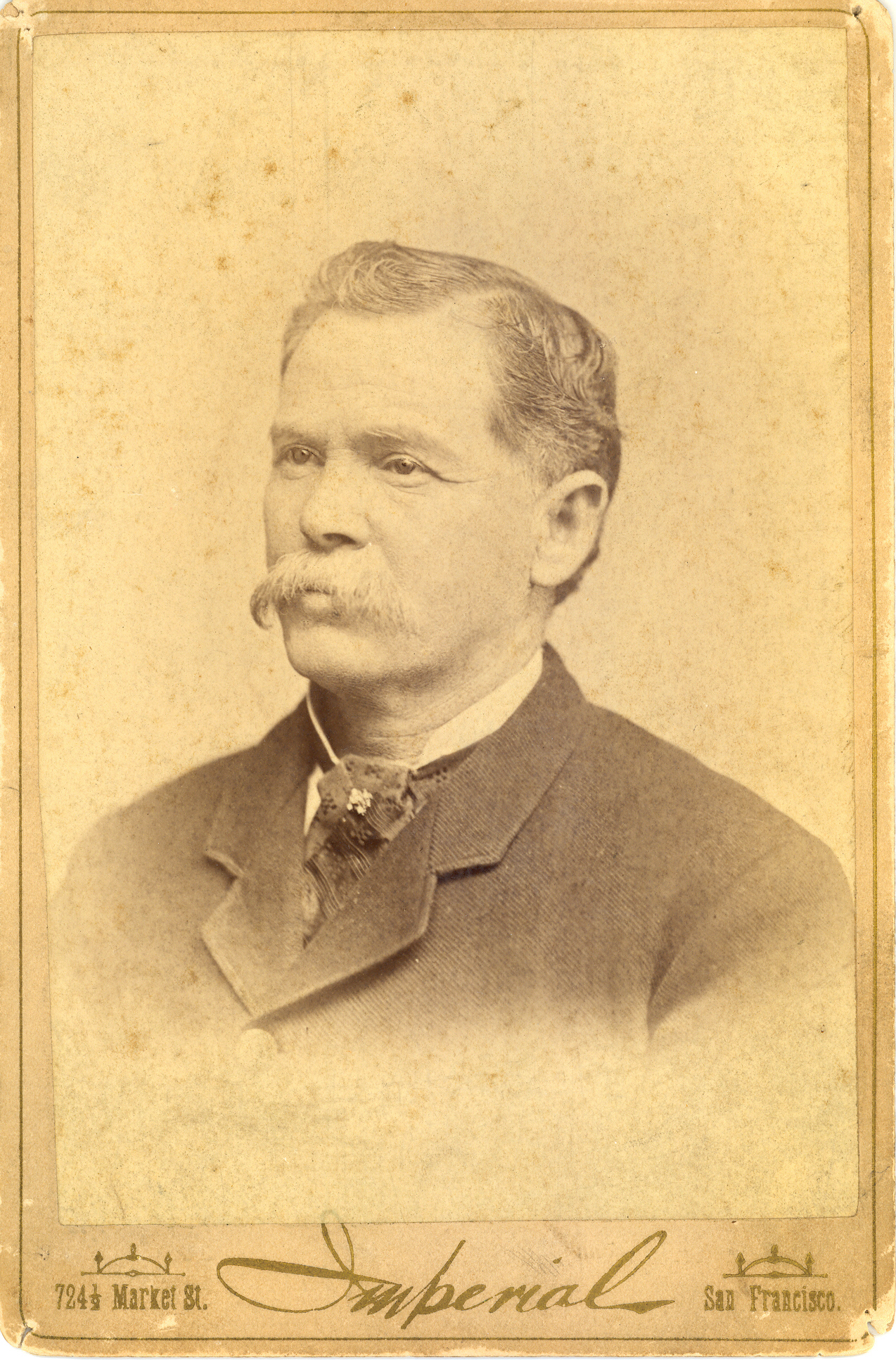 A man with a grey mustache wearing a black suit and tie with a tie pin in center. Black and white portrait.