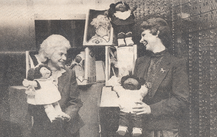 Two women in a bank vault with safe deposit boxes. The two women stand holding Cabbage Patch dolls in front of a pile of boxes for more dolls.