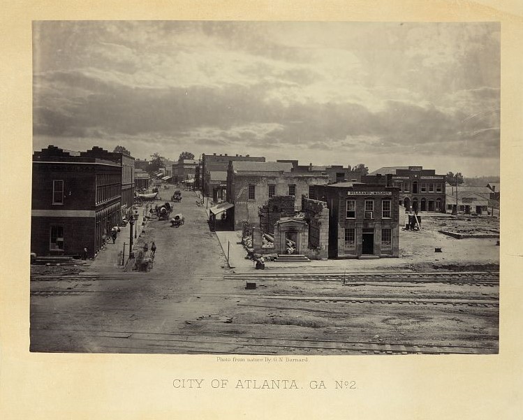 Black and white photograph on yellowing cardboard. The picture shows the destruction of Atlanta during the Civil War with many buildings in ruins and the railroad tracks damaged.