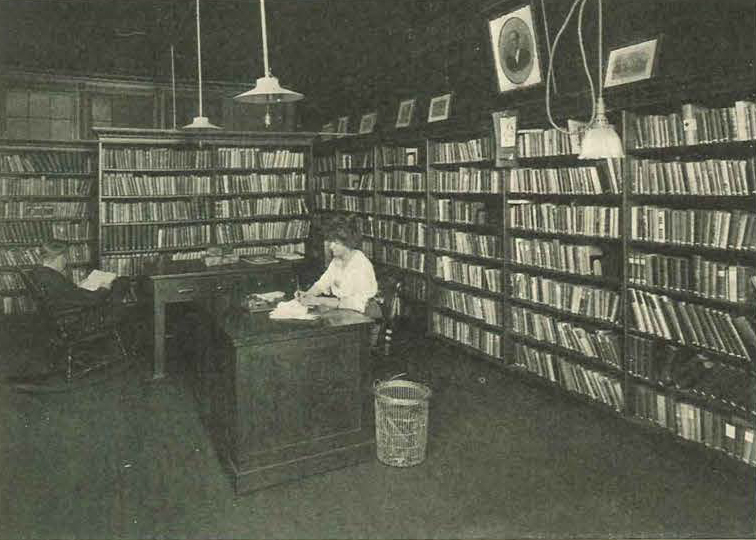 Woman sitting behind a desk inside a library room lined with shelves of books. Black and white image.