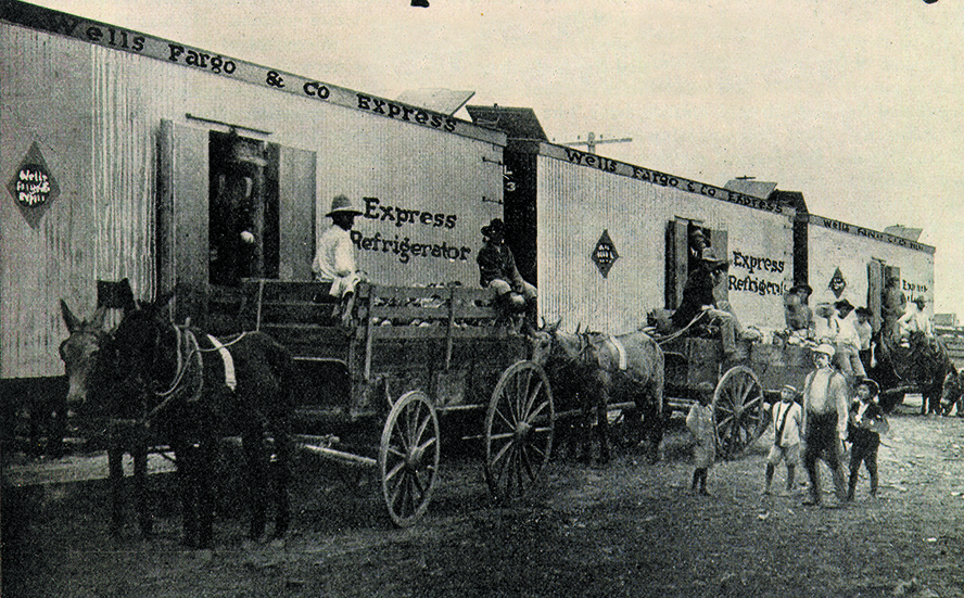 Wagons filled with produce pulled by mules or donkeys. The three wagons line up along three rail cars with lettering that reads: Wells Fargo and Co. Express, Express Refrigerator. Black and white image.