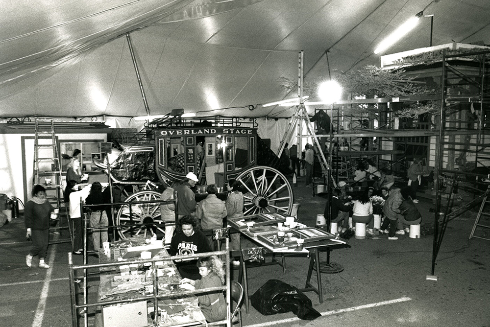 Black and white picture taken inside a large tent. A group of people gather around a stagecoach. Ladders, tables, and other construction materials are visible throughout the tent.