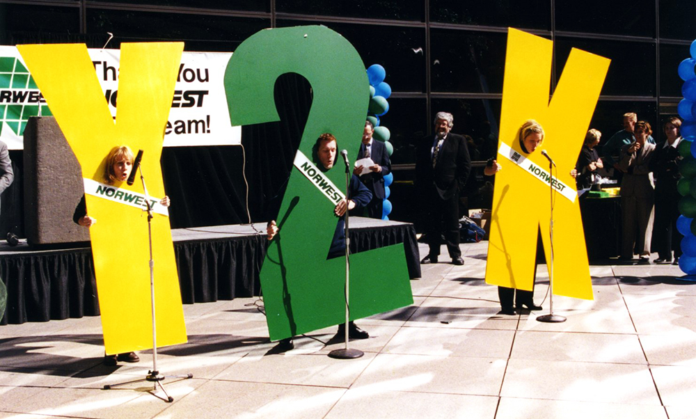 Three people on a stage wearing costumes that spell out Y2K. The Y and K are yellow. The 2 is green. Each person stands in front of a microphone. A Norwest sign is visible in the background.