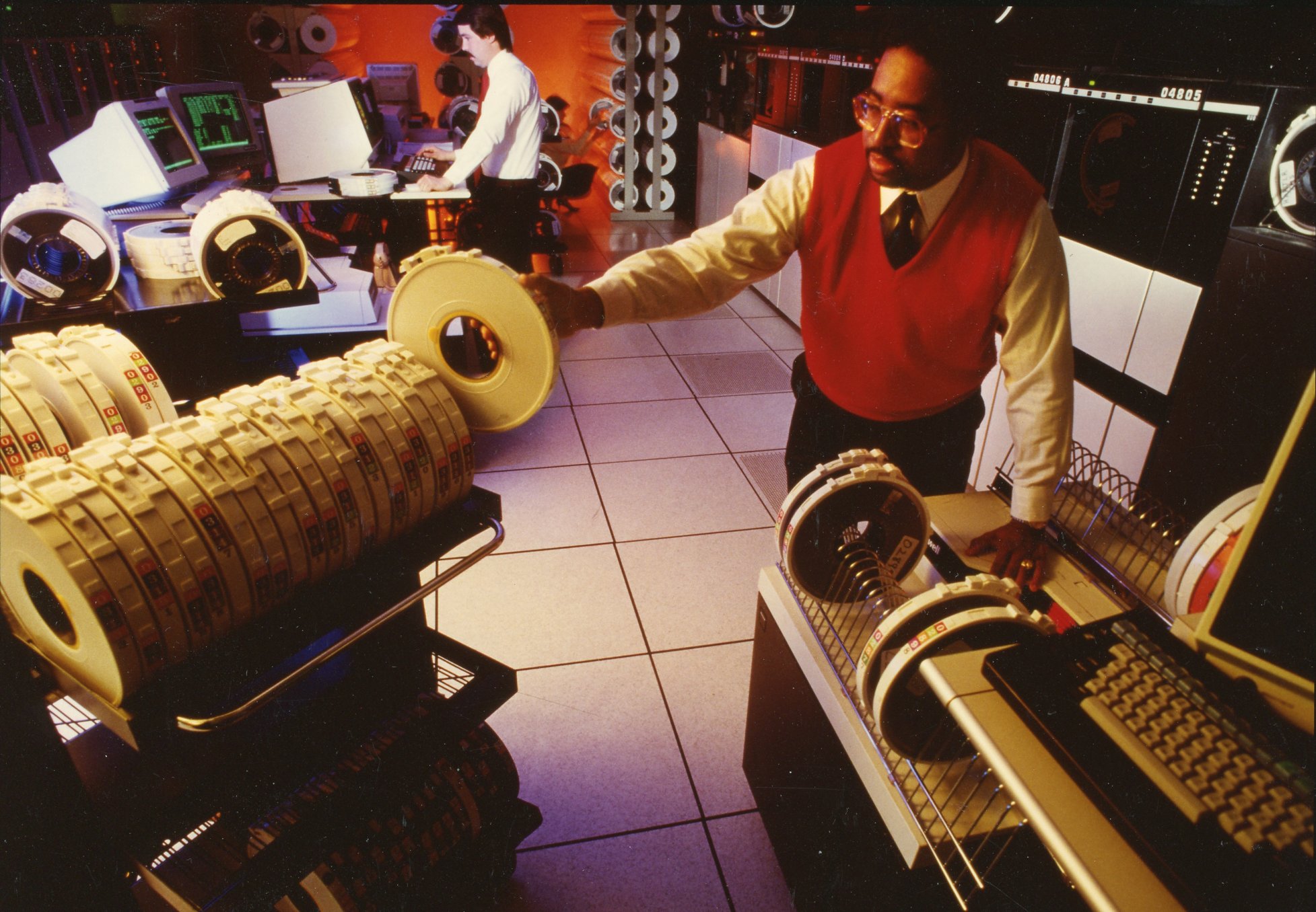 Man in red vest holds a magnetic tape wheel. In the background are large computers and a man accessing a computer monitor. 