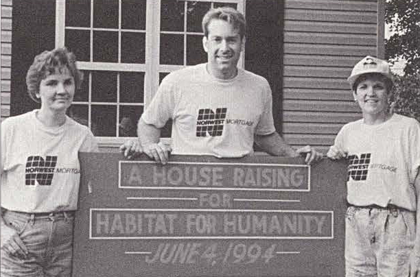 Three people in Norwest Mortgage t-shirts stand behind sign that reads: A house raising for Habitat for Humanity June 4, 1994. In the background is a newly built house. 