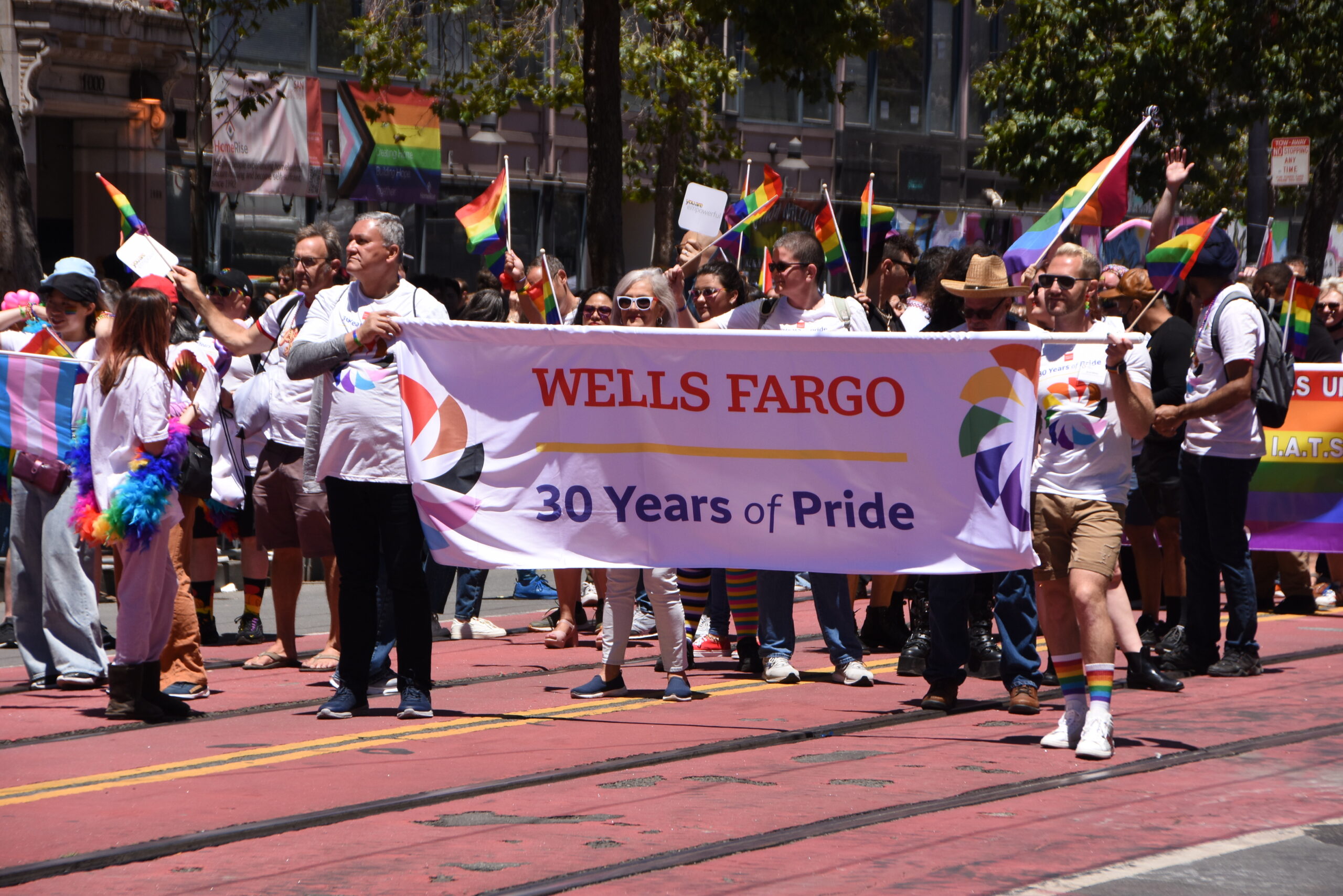 Group of Wells Fargo employees carry a banner that reads Wells Fargo 30 years of pride down a busy street with crowds on the side. 