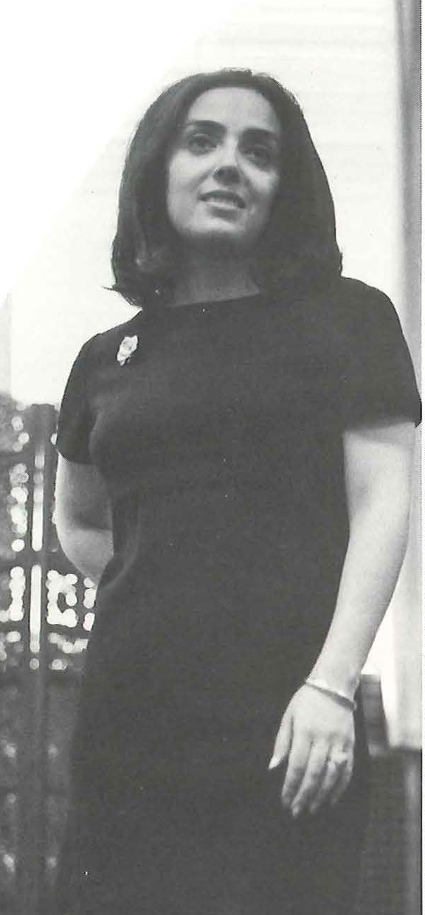 A woman in a black dress with a pin on her lapel stands with one hand behind her back. Image link will enlarge image.