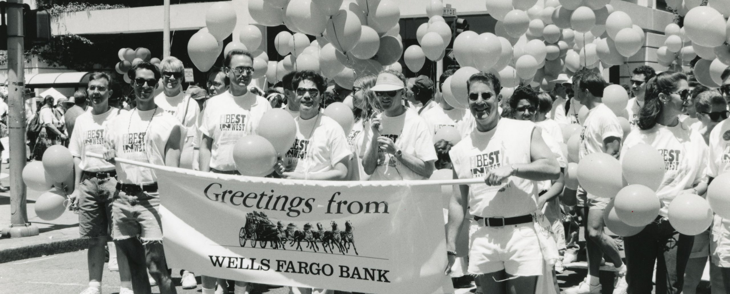 A group of people carrying a Wells Fargo banner with stagecoach image while marching in a Pride parade. In the background is a large building and balloons.
