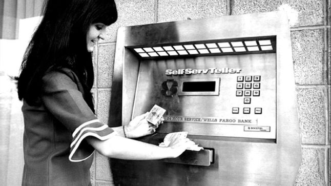 A woman removes cash from a slot on a wall mounted ATM machine with her right hand. Her left hand holds an ATM card. The machine has multiple buttons and reads Self Serv Teller. Image link will enlarge image.