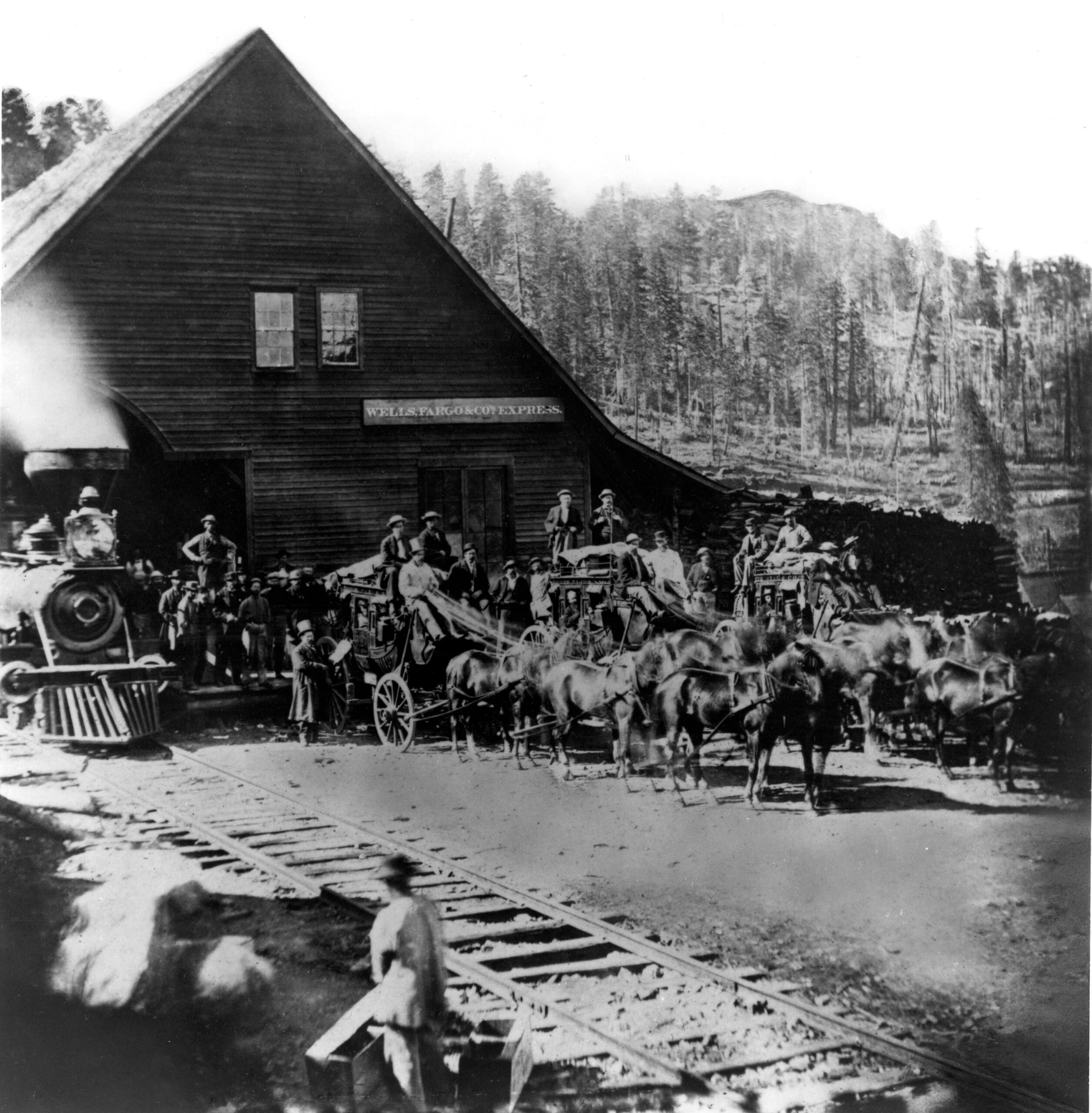 Three stagecoaches with horses lined up outside wooden building with Wells Fargo sign. A steam train sits on tracks next to the building. A crowd of people gather and sit on the coaches.