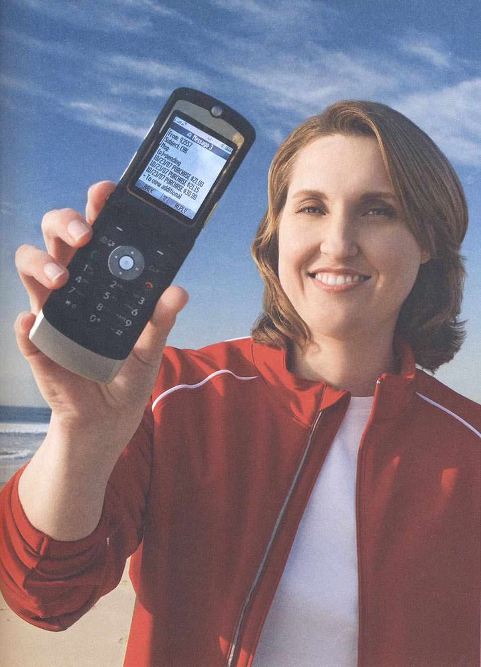 Smiling woman in red jacket on a beach holds up a flip phone that has a series of financial related text messages. 