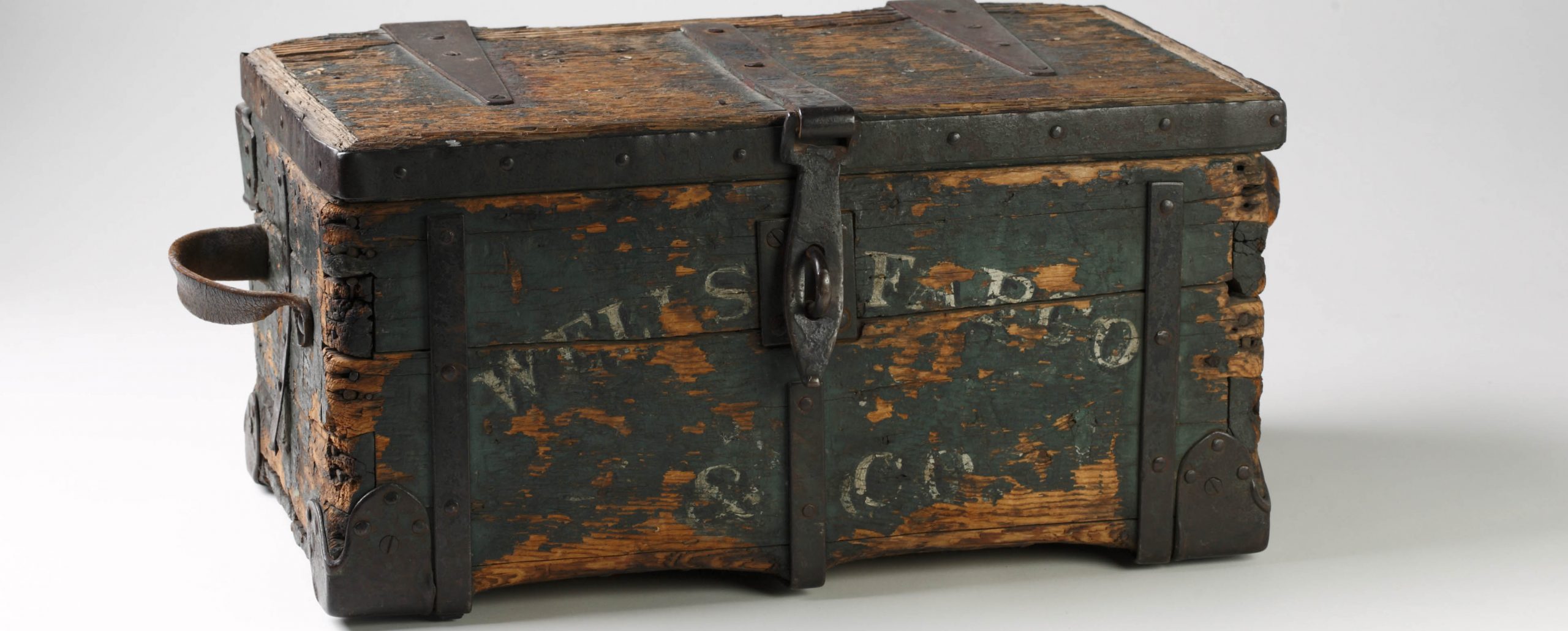 A rectangular wood box with a leather strap handle on left and Wells Fargo & Co on the front. Years of use have stripped away the green paint, but some is still visible on front panel.