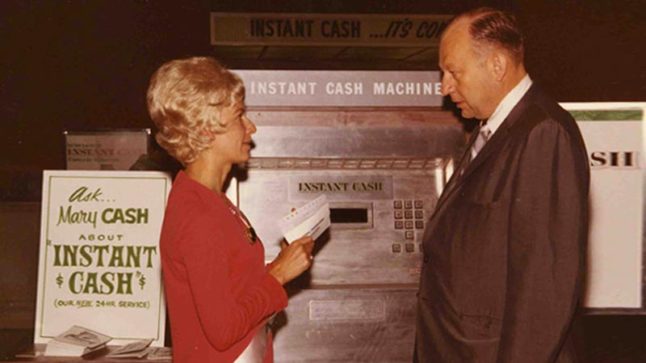 A woman in a red dress and a man in a grey suit stand before an Instant Cash Machine. Signage to the left reads: Ask Mary Cash About Instant Cash. Image link will enlarge image.