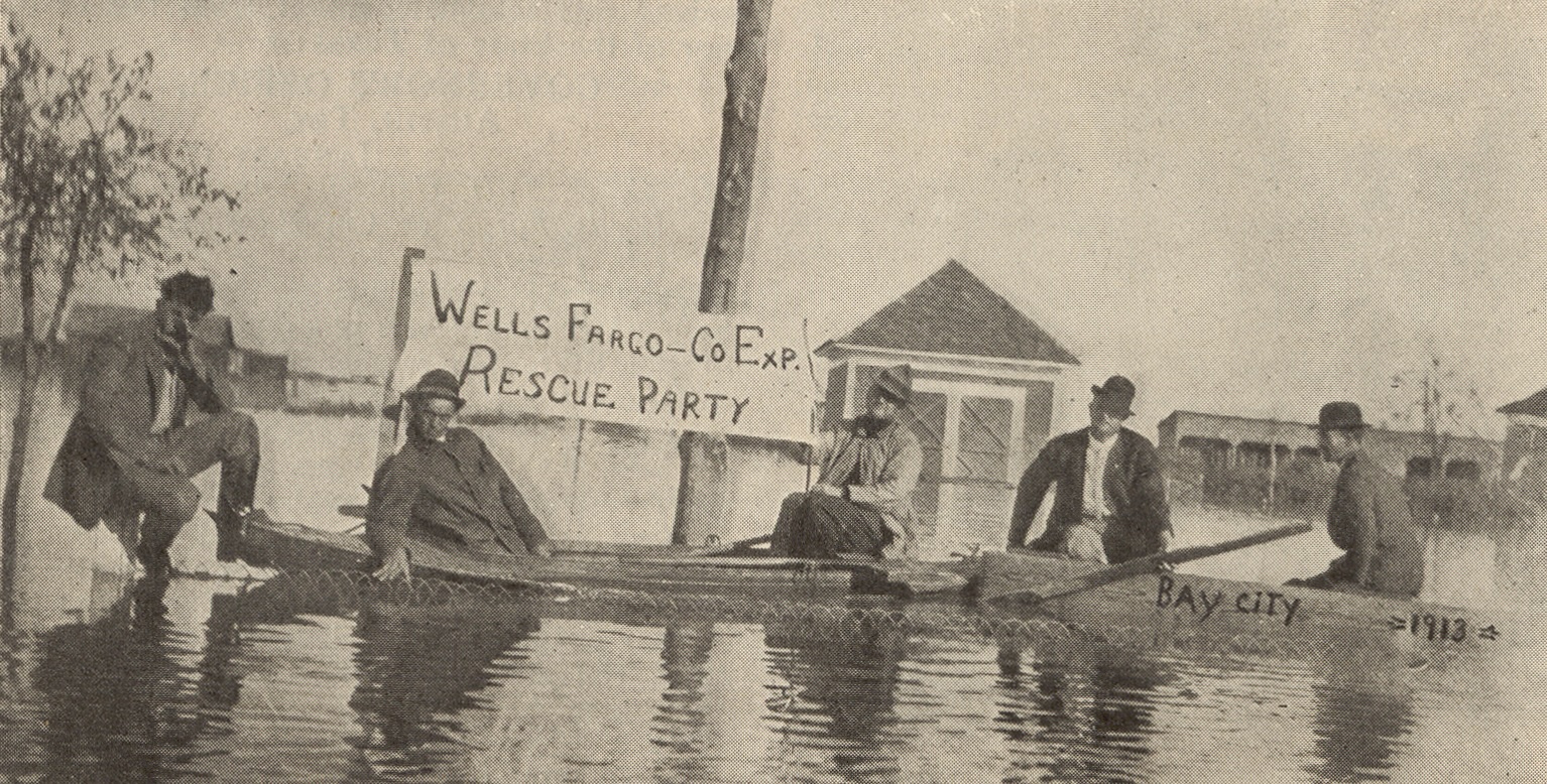 Five people in rowboats and floats with Wells Fargo Co Exp Rescue Party banner. Behind them is a farm and field underwater. A handwritten note dates the photograph to Bay City 1913. 