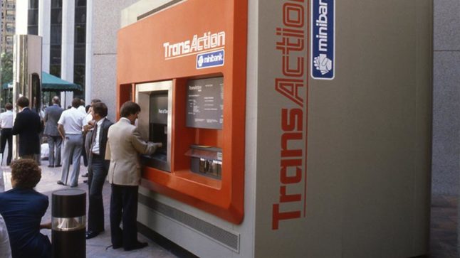 A large ATM machine on a city street is being used by a man while another man waits in line. The machine is decorated in orange and grey and reads TransAction. Image link will enlarge image.
