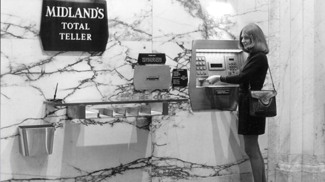 An ATM machine is mounted to a marbled wall with a sign that reads: Midland’s Total Teller. A woman stands before the machine grabbing her money as it is dispensed. Additional banking items are mounted to the wall including a trash can, a shelf holding paper and a pen. Image link will enlarge image.