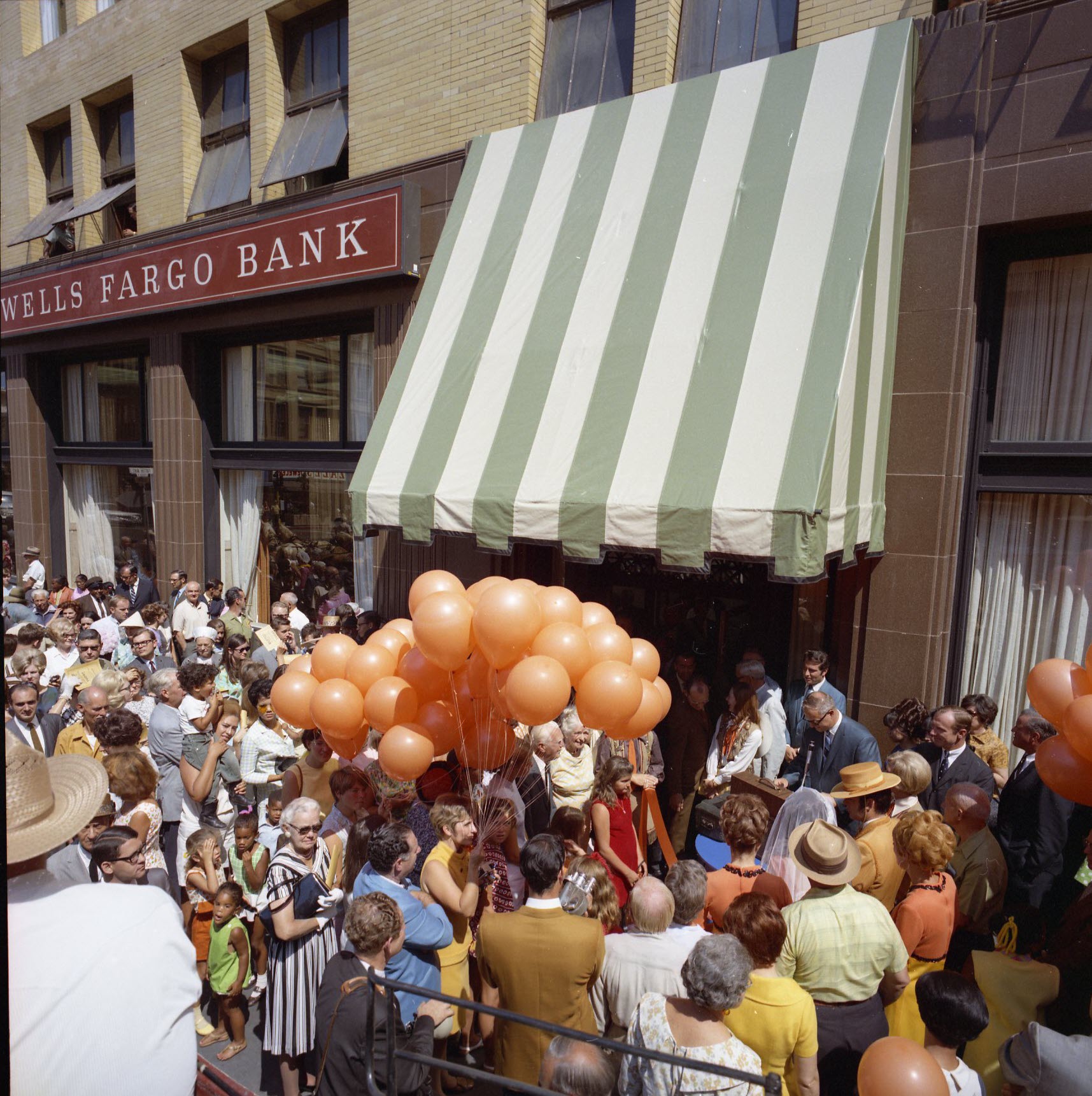 Crowd outside of a building with a Wells Fargo sign. A bouquet of orange balloons is held by a woman in a yellow dress. A man in a suit speaks at a podium. 