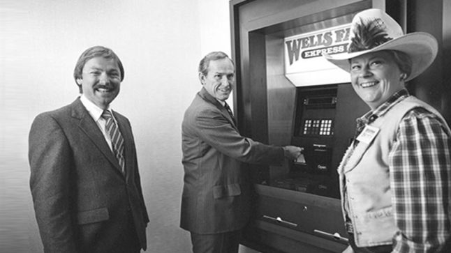 Two men in suits and a woman in a western outfit and cowboy hat stand before a wall mounted ATM machine. Image link will enlarge image.