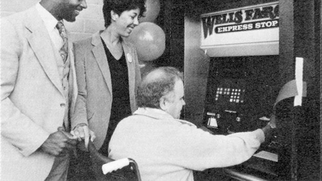 A man and a woman stand beside a man in a wheelchair as he uses a wall mounted ATM machine designed for accessibility. Image link will enlarge image.