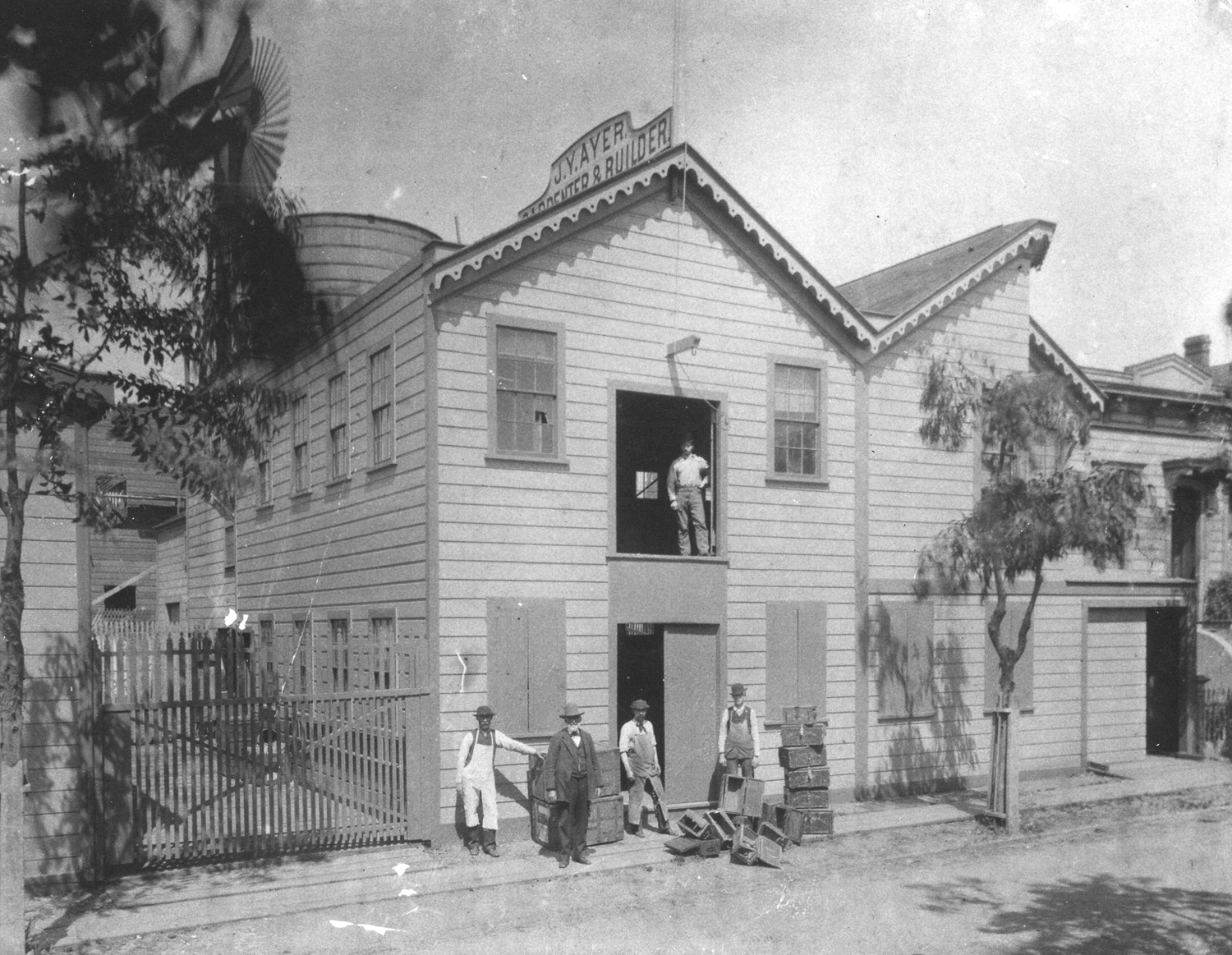 A black and white photo of two-story workshop building. Several men stand outside surrounded by wood boxes. One man stands in open loft door above. The sign on top of the building reads J.Y. Ayer Carpenter &Builder.