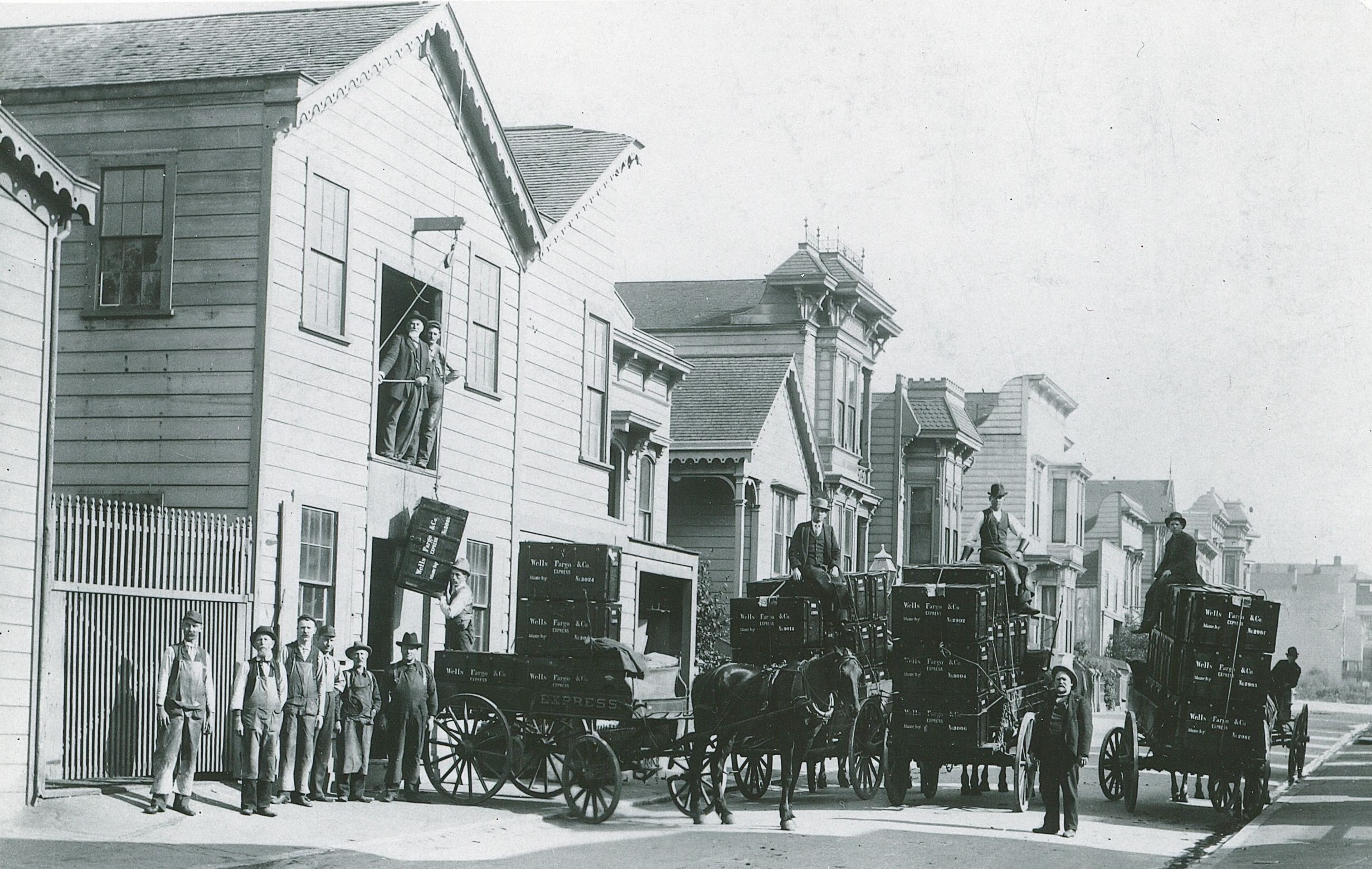 Four Wells Fargo wagons fill the street outside of a carpentry workshop building. Three are loaded with a dozen large wood trunks each. Workmen lower a trunk by rope onto a fourth wagon. Workers and wagon drivers stand on and around the wagons.