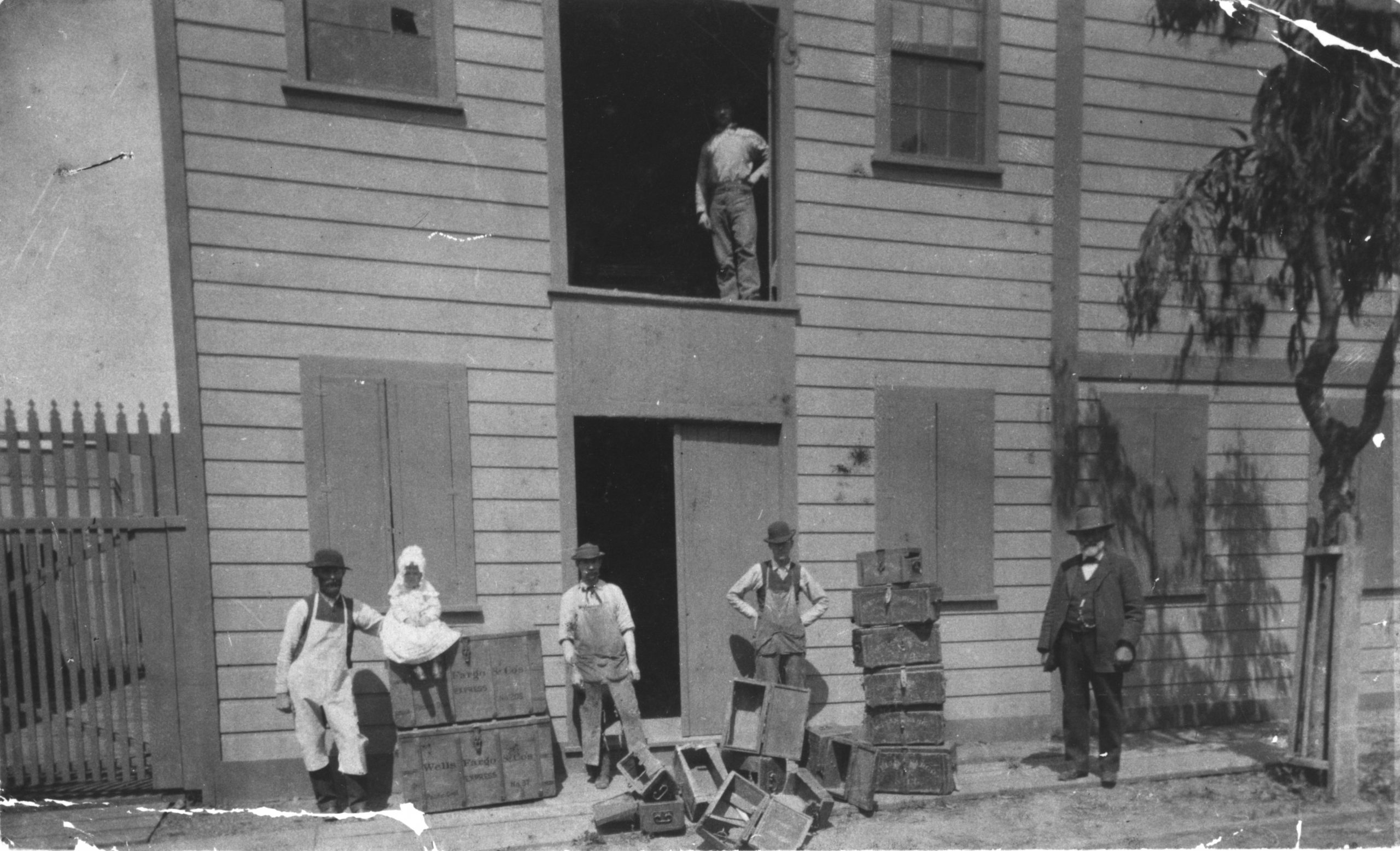A black and white photo of building front shows five men in work clothes and stacks of wood boxes. A little girl sits on top of one stack of boxes. The older, bearded man at right is J.Y. Ayer.