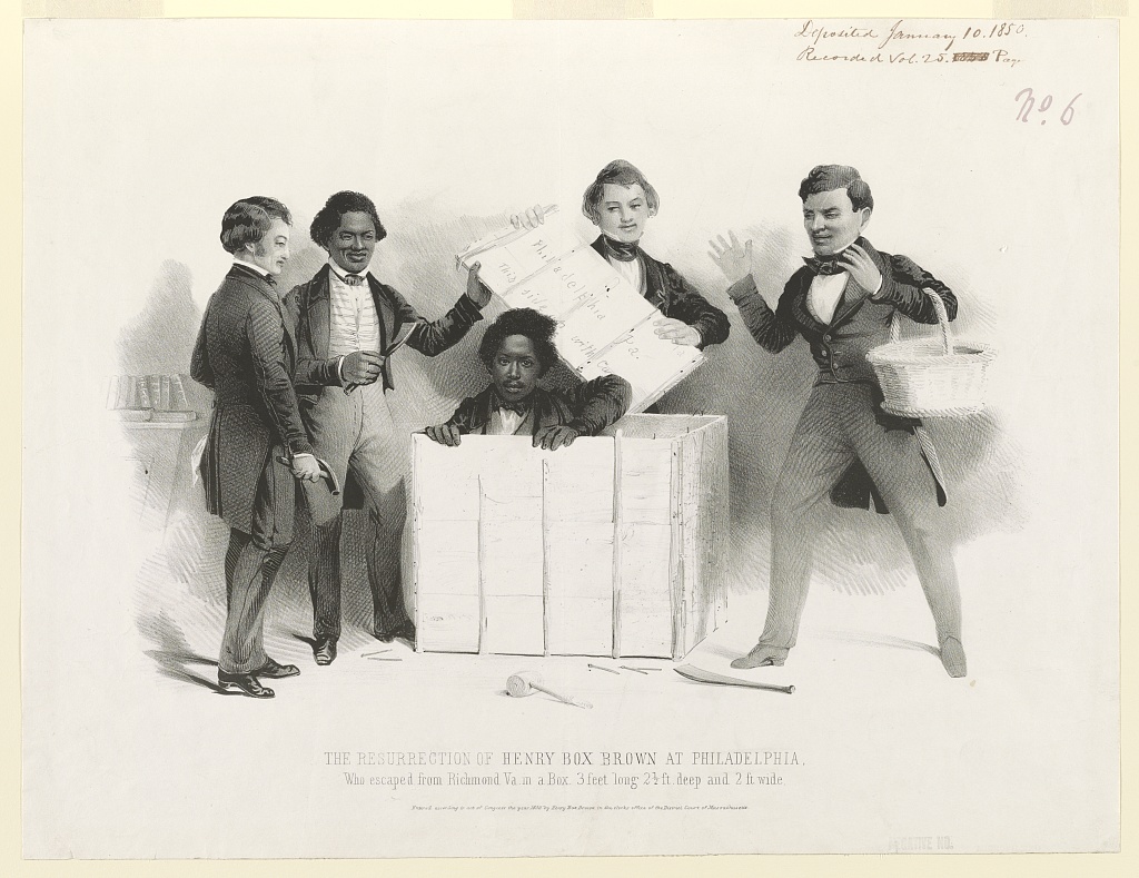 A black and white image shows four men in suits standing around an open wooden box with a man inside who has his head, shoulders, arms, and hands showing, as he emerges from it.