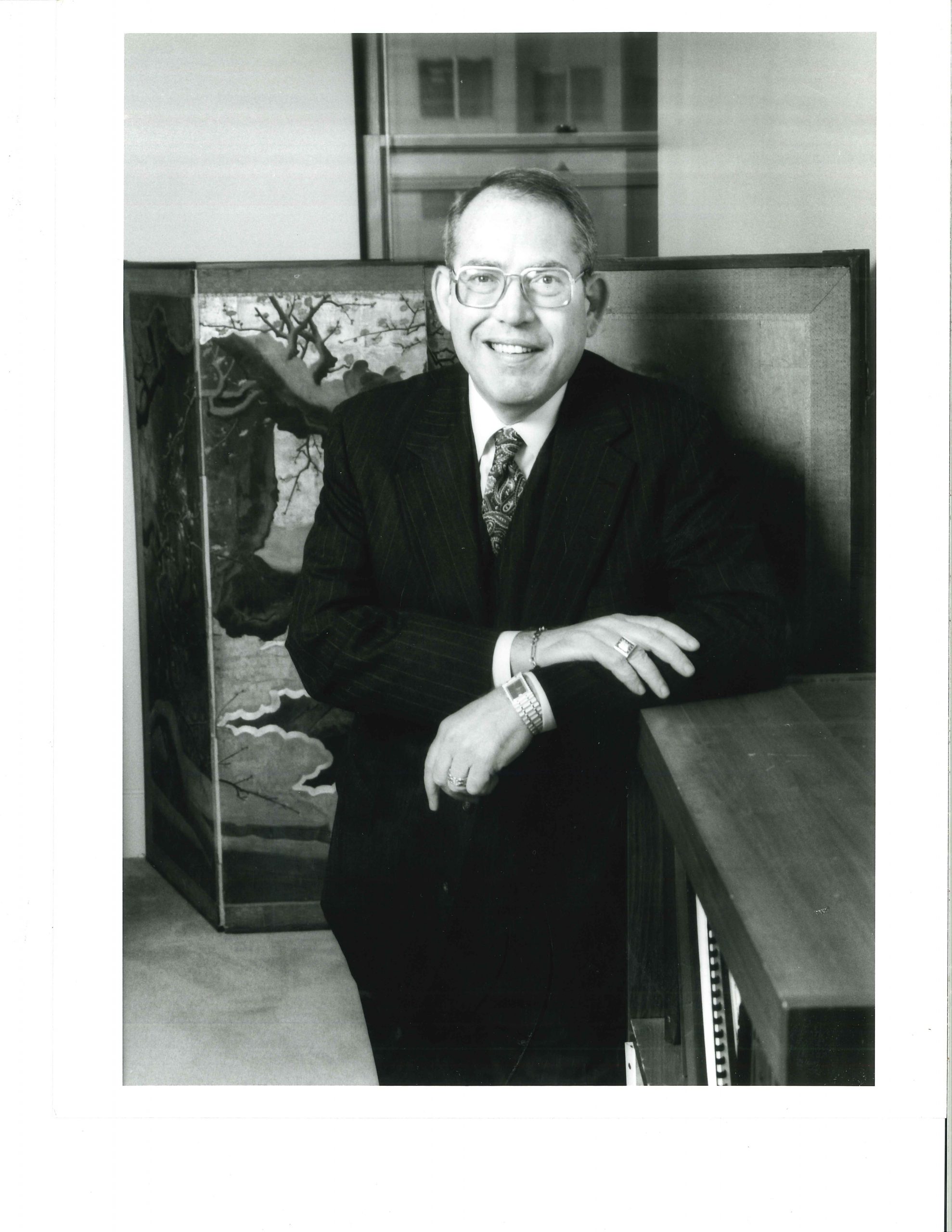 Douglas P. Holloway, in stripped suit with glasses, smiles at camera while standing in office.