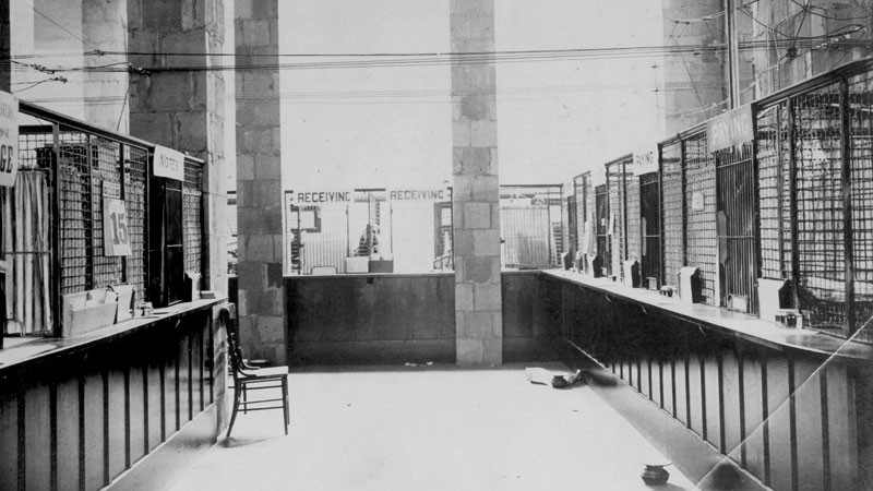 A room with bank teller cages on three sides. Large windows let in light in the background. A single chair sits in the customer area. Image link will enlarge image.