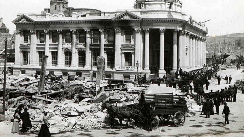 A damaged building with a large dome flying an American flag sits above a street full of rubble. A horse and wagon are among the crowd of people gathered. Image link will enlarge image.