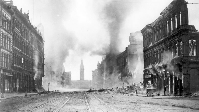 A street view with multiple buildings on fire. Huge plumes of black smoke fill the background. Black and white image. Image link will enlarge image.