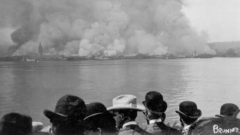 A huge mass of smoke covers the city in the background while evacuees on a boat watch in the foreground. The bay separates the two. Image link will enlarge image.