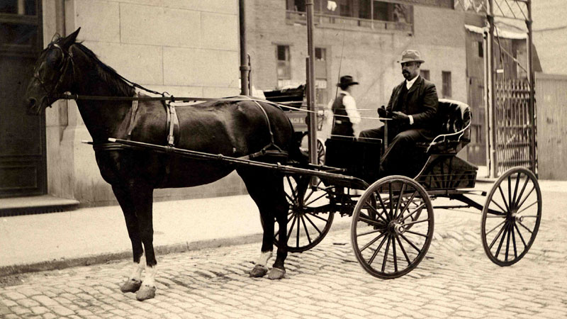 A horse and buggy parked on a cobbled street with a building in the background. The driver is wearing a black suit and hat. Image link will enlarge image.