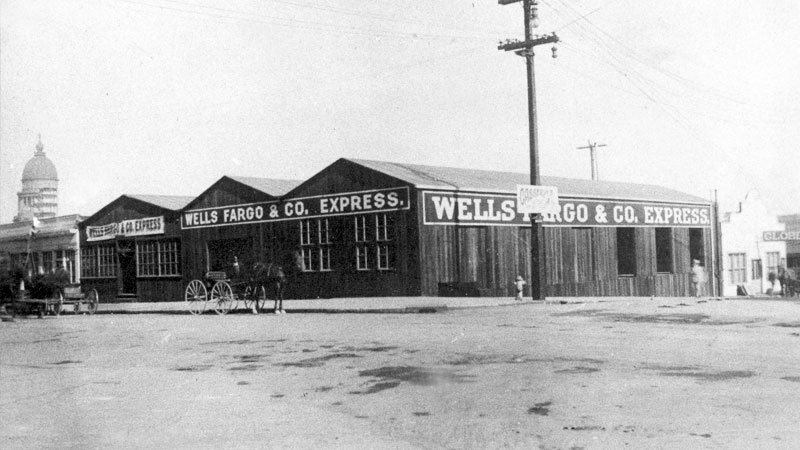 A street view of a row of wood buildings. Wells Fargo & Co Express is painted across the front and sides of the buildings. A horse and buggy drive past on the street. Image link will enlarge image.