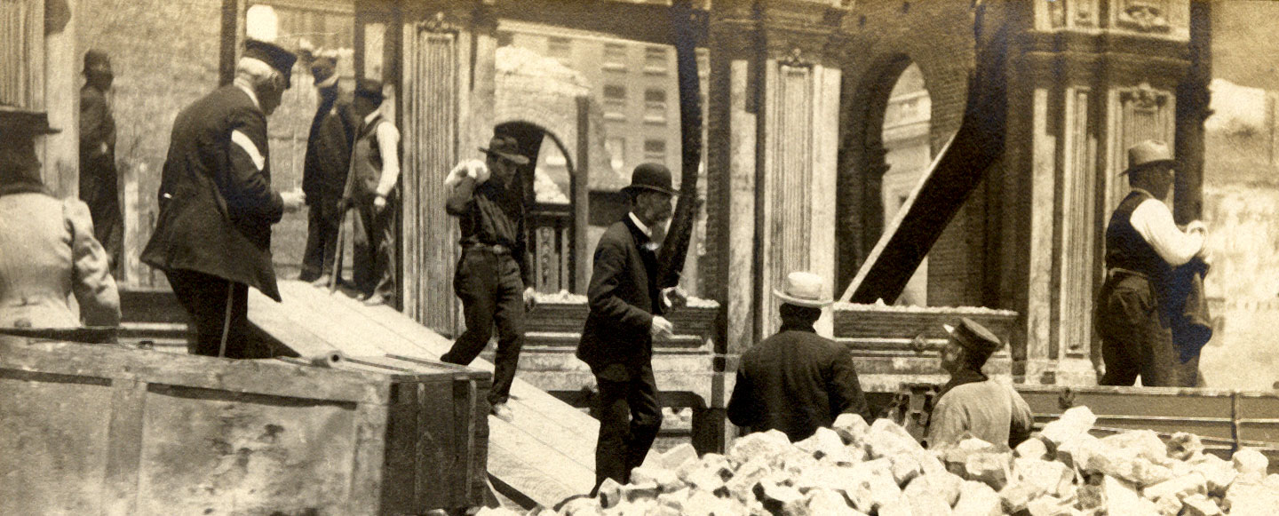People sort through the rubble of a building destroyed by an earthquake. The foreground is full of pieces of brick and a dumpster. Image in black and white.