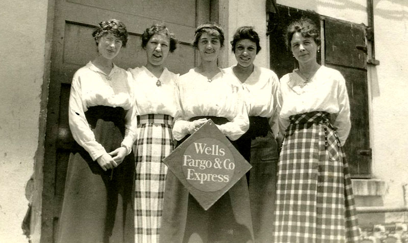 Five women wearing long skirts and white blouses stand outside of a building doorway. The middle woman holds a Wells Fargo & Co. diamond sign. Image link will enlarge image.