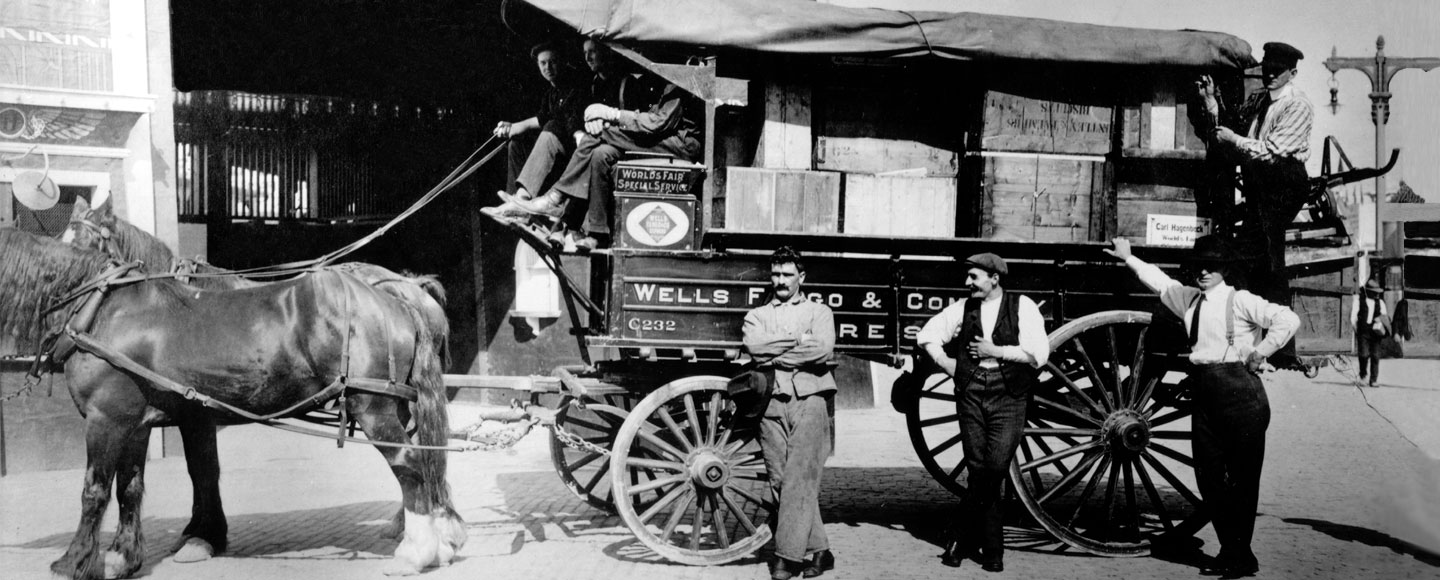 A loaded Wells Fargo wagon pulled by two horses. Three men stand in front of the wagon, one man stands on the rear bumper and three men are seated in the driver’s seat. Image in black and white.