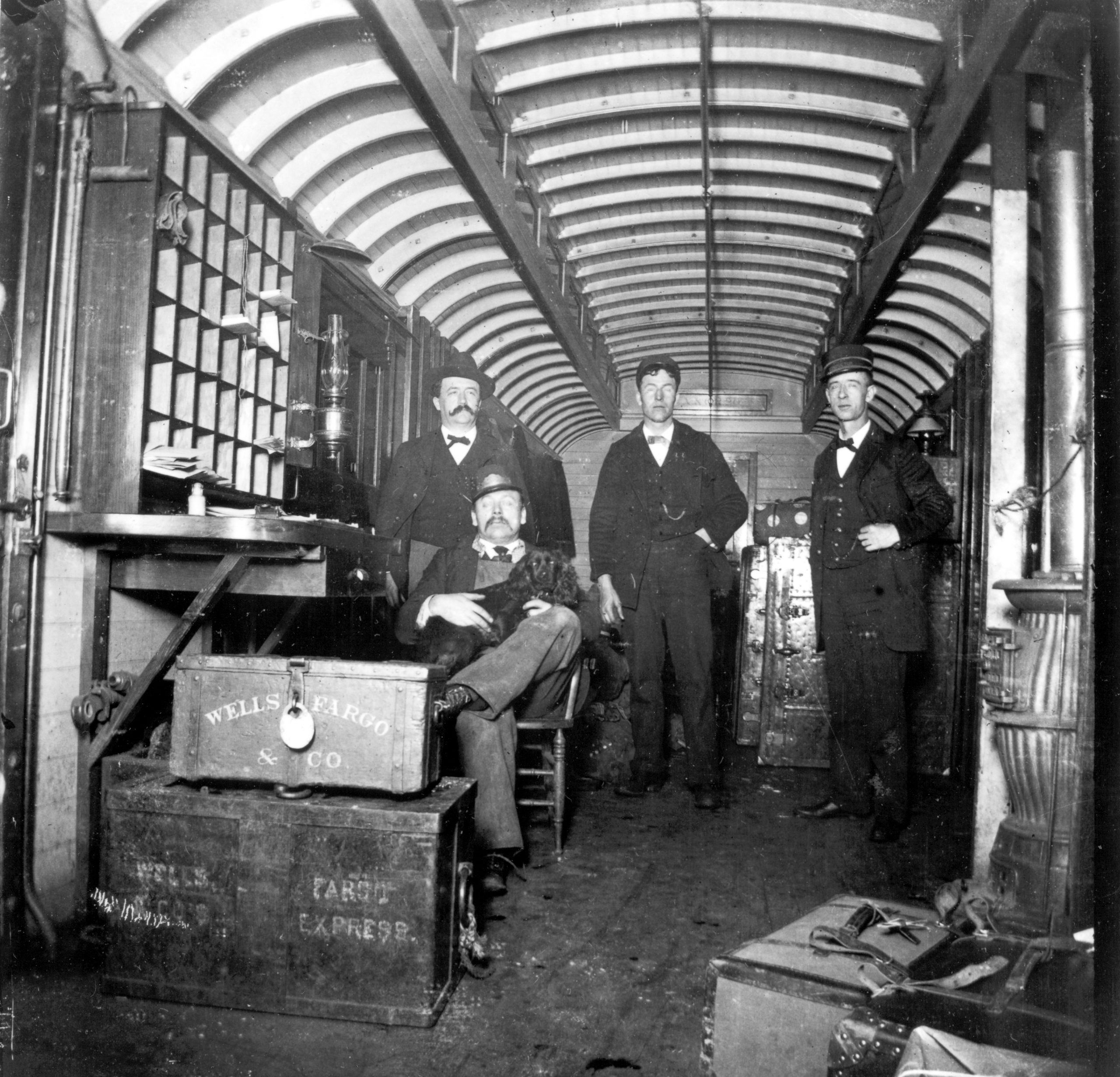 The interior of a wood railroad car shows three men standing and one sitting in chair with a small black dog on his lap. In the foreground are various trunks and locked boxes labeled Wells Fargo & Co.