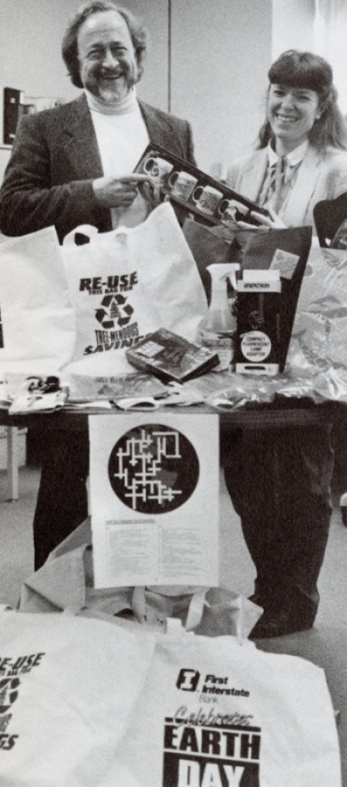 Two people at table displaying First Interstate Bank of Oregon’s Earth Day tote bags, mugs, and other promotional items.