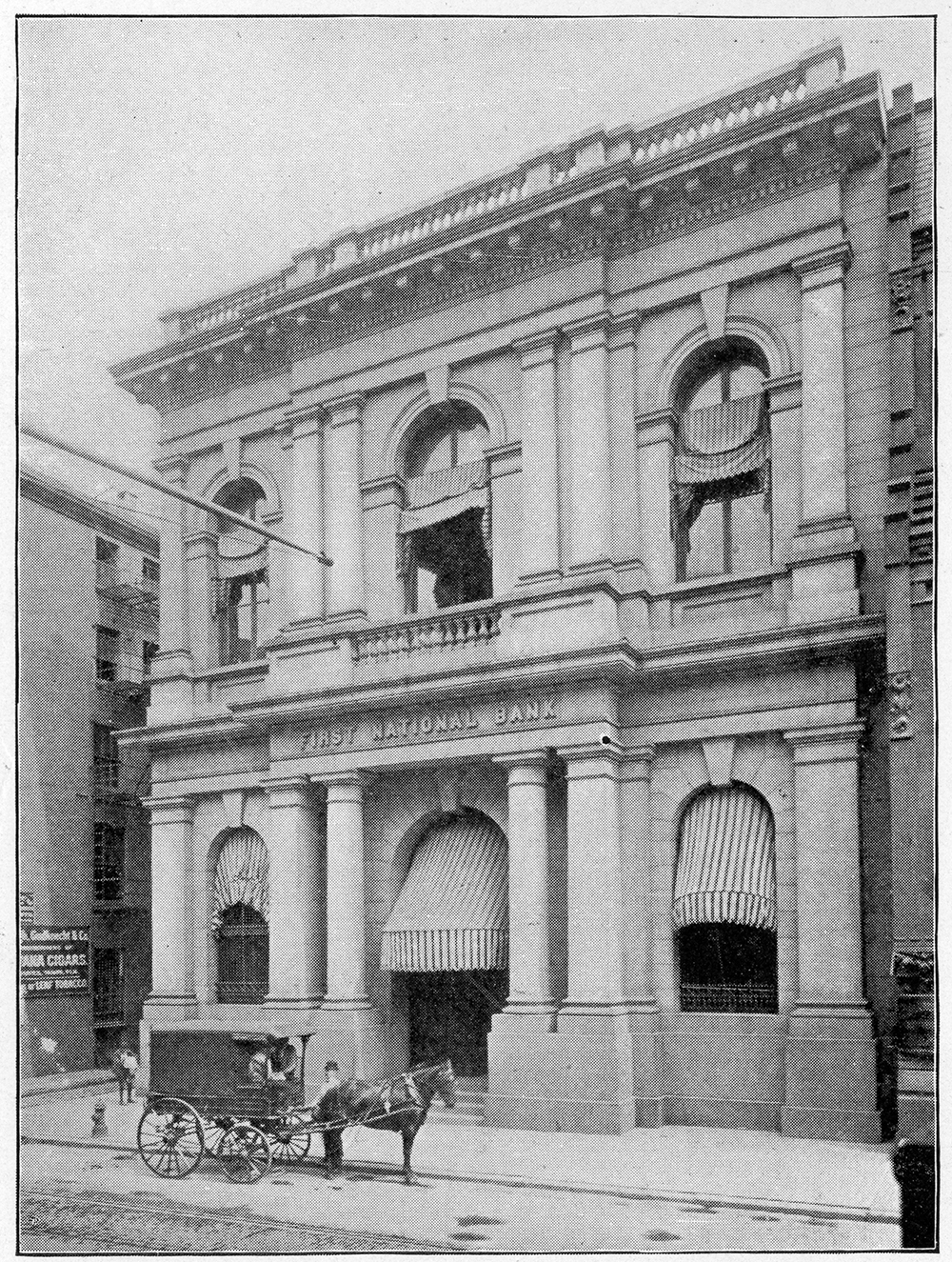 A two-story building with arched windows and entrance. A horse and wagon pass by in front of the building.