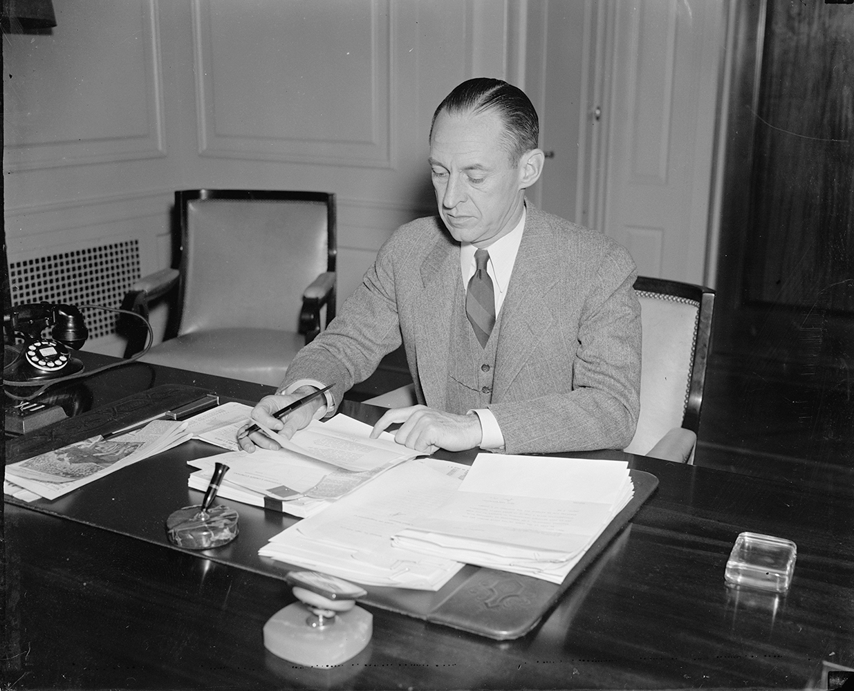 A man in a grey suit and stripped tie sits at a desk signing a large pile of papers with an ink pen. Also visible on the desk is a rotary phone and several paperweights. Image link will enlarge image.