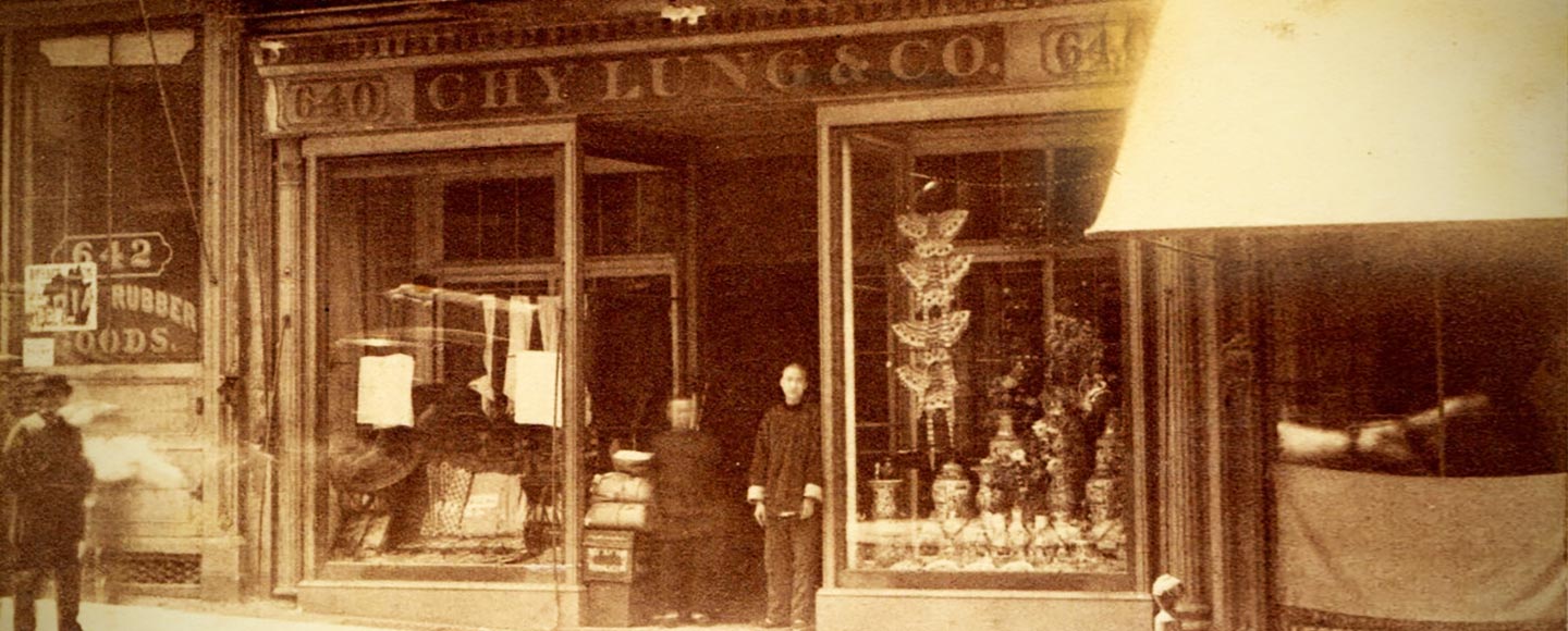 The storefront of a Chinese run business. A sign reading Chy Lung & Co.’s hangs above the doorway where two men are standing. Image in black and white.