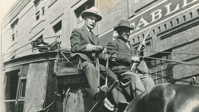 A black and white image shows two men sitting on a stagecoach, and the back of a horse in front of them. The man on the left has the reins, and the man on the right holds a gun. Behind them is a building with part of the word stable seen.