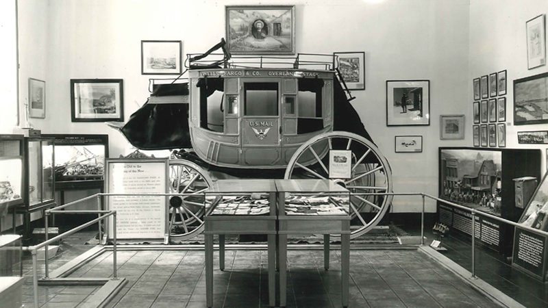 A black and white image shows a stagecoach in front of a wall with pictures. Nearby are some tables with items on them and other frames on the wall.